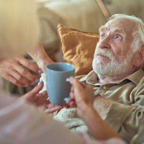 Elderly gray-haired man sick at home while his wife caring for him and holding hot drink for him. Care and health concept